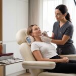 Clinician marking a patient's jawline before filler treatment in a clean aesthetic clinic; patient reclined with a cool compress at the chin.